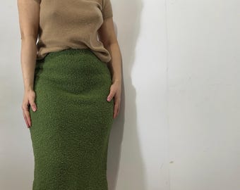 May include: A short-sleeved, tan-colored sweater paired with a textured, olive-green pencil skirt. The skirt reaches the knees. The outfit is worn by a person standing against a white wall.