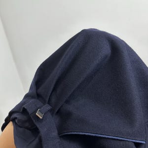 May include: A dark navy blue wool-like fabric garment featuring a structured hood with a small fabric loop and a metallic silver-toned rectangular buckle. The piece includes a deep pocket with a thin blue trim and a smooth, dark navy blue inner lining.