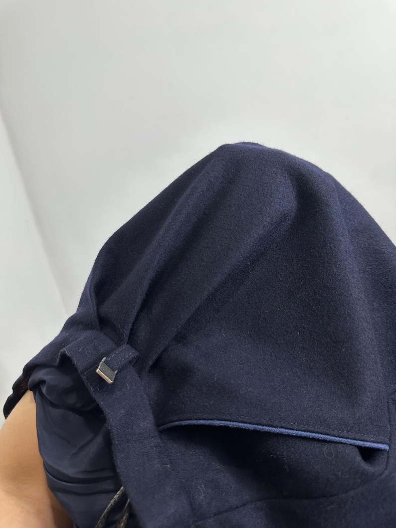 May include: A dark navy blue wool-like fabric garment featuring a structured hood with a small fabric loop and a metallic silver-toned rectangular buckle. The piece includes a deep pocket with a thin blue trim and a smooth, dark navy blue inner lining.