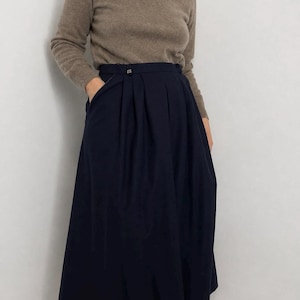May include: A taupe long-sleeve turtleneck sweater paired with a high-waisted navy blue midi skirt. The skirt features deep vertical pleats, a structured waistband with a small metallic button detail, and side pockets, falling below the knees.