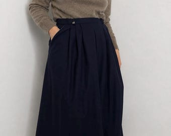 May include: A taupe long-sleeve turtleneck sweater paired with a high-waisted navy blue midi skirt. The skirt features deep vertical pleats, a structured waistband with a small metallic button detail, and side pockets, falling below the knees.