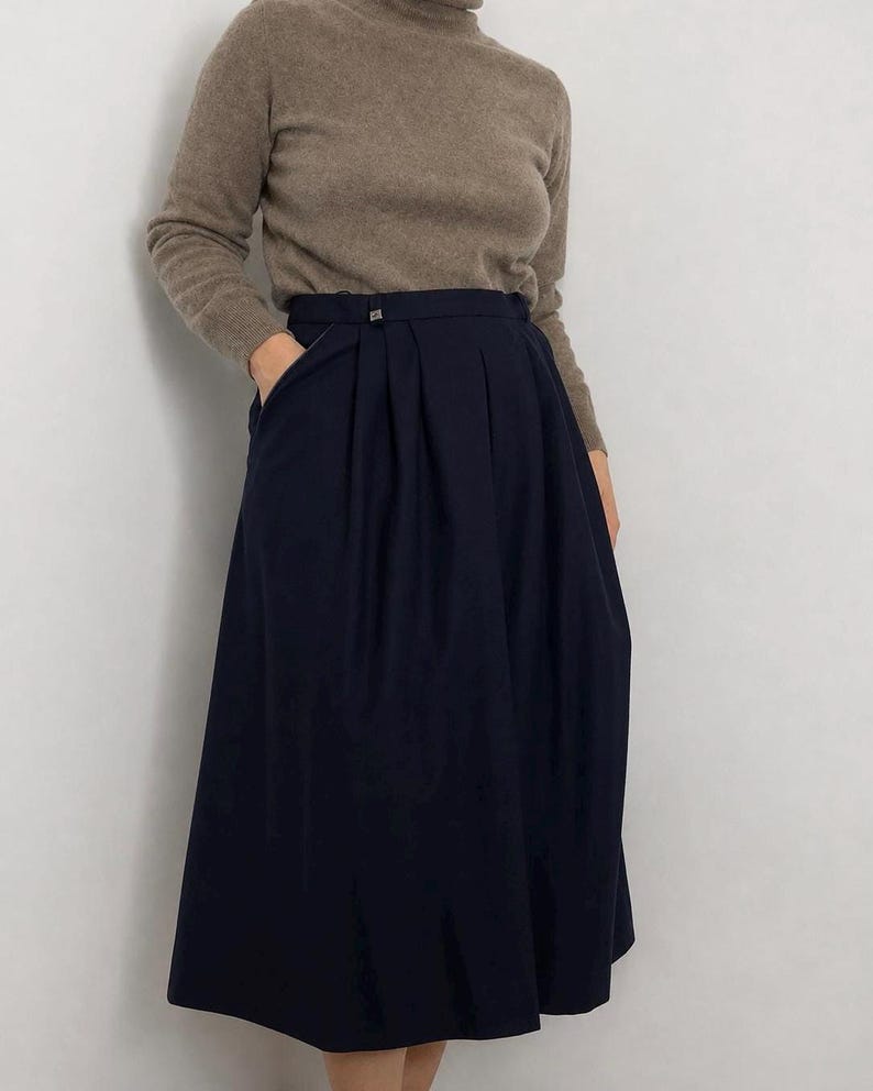May include: A taupe long-sleeve turtleneck sweater paired with a high-waisted navy blue midi skirt. The skirt features deep vertical pleats, a structured waistband with a small metallic button detail, and side pockets, falling below the knees.