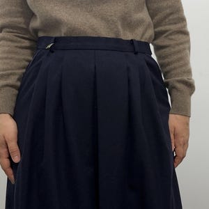 May include: A high-waisted navy blue skirt featuring deep vertical pleats, a structured waistband with belt loops, and a small metallic accent on the left side, paired with a long-sleeved beige sweater with a ribbed crew neckline and cuffs.