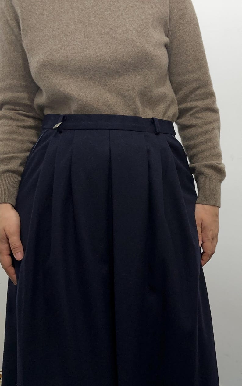 May include: A high-waisted navy blue skirt featuring deep vertical pleats, a structured waistband with belt loops, and a small metallic accent on the left side, paired with a long-sleeved beige sweater with a ribbed crew neckline and cuffs.