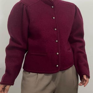 May include: A deep burgundy wool jacket featuring a high mandarin collar, long sleeves with gathered shoulders, and a front button closure with small, round, metallic silver buttons. The jacket includes two horizontal welt pockets at the waist.
