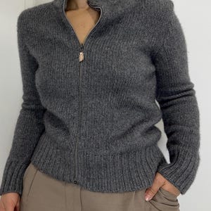 May include: A charcoal gray zip-up sweater with a ribbed texture. The sweater has a high collar and long sleeves. The zipper pull is a light gold color. The sweater is paired with tan pants.