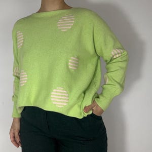 Puede incluir: Un suéter verde claro con grandes círculos a rayas rosas y blancas. El suéter de manga larga tiene cuello redondo y está hecho de un material suave. El suéter se combina con pantalones verde oscuro.