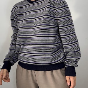 May include: A long-sleeve knit sweater featuring a horizontal striped pattern in shades of grey, black, and purple. The garment has a solid dark navy blue crew neckline, matching ribbed cuffs at the wrists, and a matching ribbed hem at the waist.