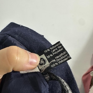 May include: A dark navy blue garment with a black fabric care label. The label displays the text 70% Schurwolle, 30% Polyester, and 100% Viskose, along with the words Hauptfutter and Doublure. A small circular logo is visible on the left side.