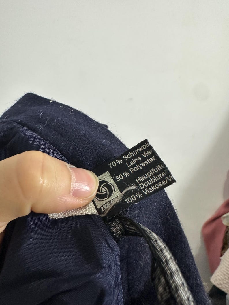 May include: A dark navy blue garment with a black fabric care label. The label displays the text 70% Schurwolle, 30% Polyester, and 100% Viskose, along with the words Hauptfutter and Doublure. A small circular logo is visible on the left side.