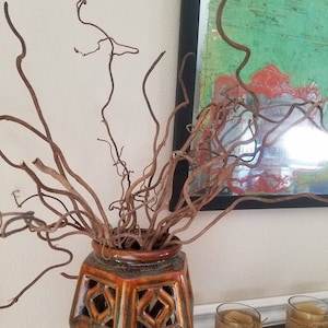 May include: A decorative ceramic vase with an orange and brown glaze, featuring a geometric cut-out design. The vase holds several dried, twisted brown branches. Two glass candle holders wrapped in burlap sit on a tray.