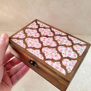 Puede incluir: Una caja rectangular de madera con un diseño floral en la tapa. La tapa presenta un patrón de flores rosas sobre un fondo blanco, enmarcado por madera marrón oscuro. La caja tiene un pequeño cierre metálico.