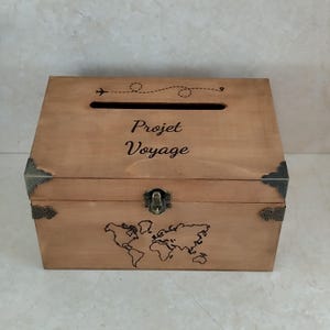 May include: A wooden box with a slot on top and a world map design on the front. The box is light brown with metal corner accents and a clasp. The text "Projet Voyage" is written on the front, along with an airplane graphic.