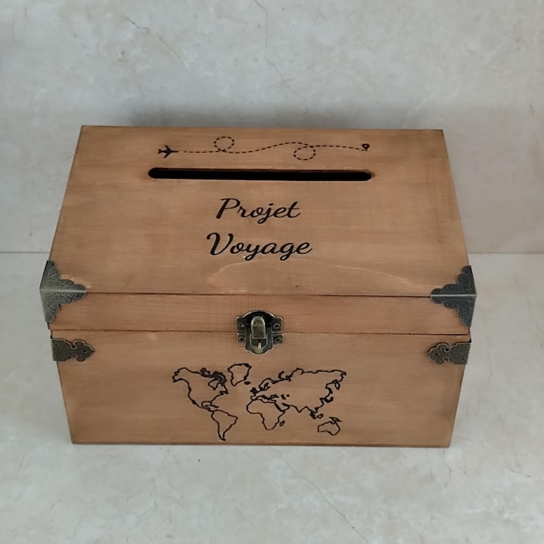 May include: A wooden box with a slot on top and a world map design on the front. The box is light brown with metal corner accents and a clasp. The text "Projet Voyage" is written on the front, along with an airplane graphic.