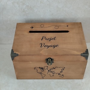 May include: A wooden box with a slot on top and a hinged front. The box is light brown with dark metal corner accents and a clasp. The words "Projet Voyage" are written on the front, along with a world map and an airplane graphic.