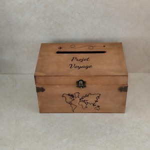 May include: A wooden travel-themed box with a slot on top and a hinged front. The box is light brown with a world map and the words "Projet Voyage" etched on the front. A small airplane graphic and dotted line are above the text.