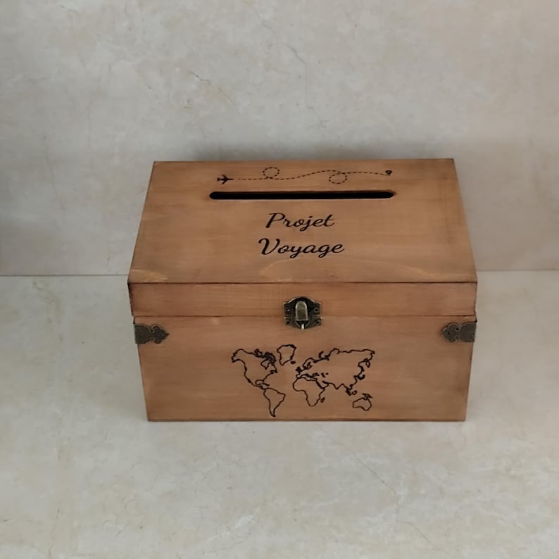 May include: A wooden travel-themed box with a slot on top and a hinged front. The box is light brown with a world map and the words "Projet Voyage" etched on the front. A small airplane graphic and dotted line are above the text.