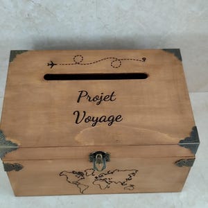 May include: A wooden travel-themed box with the words "Projet Voyage" and a world map design. The box has a slot on top and metal corner accents. A small airplane graphic and dotted line are above the text.