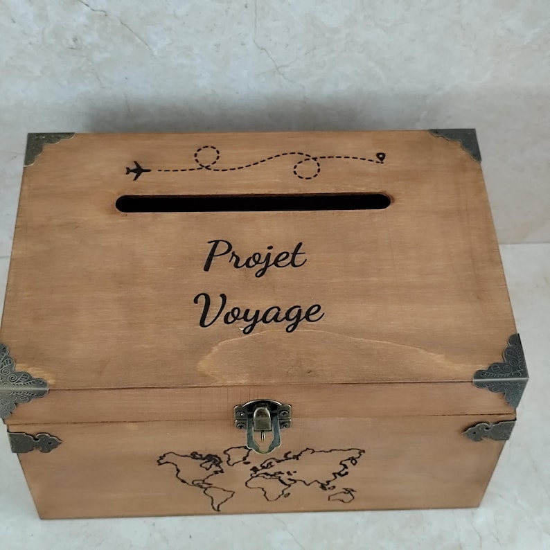 May include: A wooden travel-themed box with the words "Projet Voyage" and a world map design. The box has a slot on top and metal corner accents. A small airplane graphic and dotted line are above the text.
