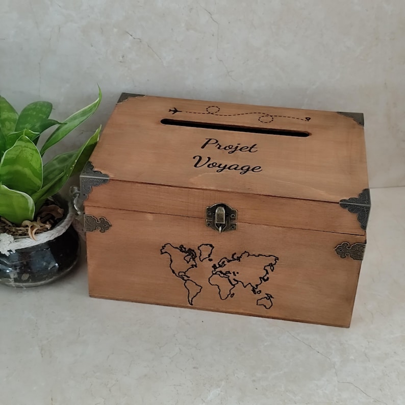 May include: A wooden travel-themed box with a slot on top and a world map design on the front. The box has metal corner protectors and a clasp. The text "Projet Voyage" is above the slot, and an airplane graphic is above the text.