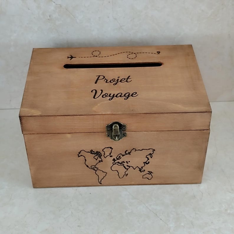 May include: A wooden box with a slot on top and a hinged front. The box is light brown with a world map and the words "Projet Voyage" etched on the front. A small airplane graphic and dotted line are above the slot.