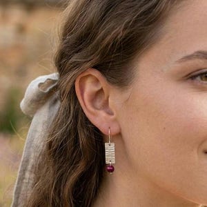 Puede incluir: Pendientes rectangulares plateados con superficie texturizada y una pequeña cuenta redonda de color rojo oscuro en la parte inferior. Los pendientes cuelgan de un gancho. El fondo está borroso.