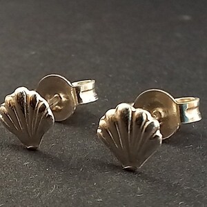 May include: A pair of small, silver seashell stud earrings. Each earring features a detailed, fan-shaped shell design. The earrings have a post and backing for secure wear, set against a dark background.