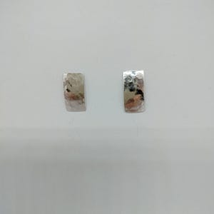 May include: Two silver rectangular earrings with a hammered texture. The earrings are displayed on a white background. The hammered texture creates a reflective surface, catching light and adding visual interest. These earrings are a simple yet elegant accessory.