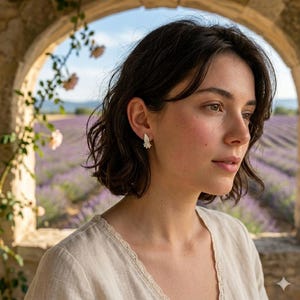 May include: A woman wearing a pair of small, delicate silver earrings. The earrings have a floral design. The woman is wearing a light-colored top. The background features a stone archway, lavender field, and a blue sky.