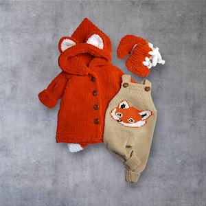 May include: A set of baby clothing in a fox theme. The set includes an orange hooded jacket with ear details, a pair of matching booties, and tan overalls with a fox face design. The items are made of a soft, textured material.