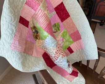 Puede incluir: Una colcha de retazos con una variedad de telas rosas, rojas, verdes y azules. La colcha está doblada y drapeada sobre una mecedora de madera.