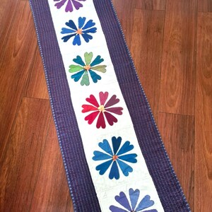 May include: A purple, blue, and green floral runner with a white background and a dark blue border. The runner features a row of colorful flowers with heart-shaped petals.