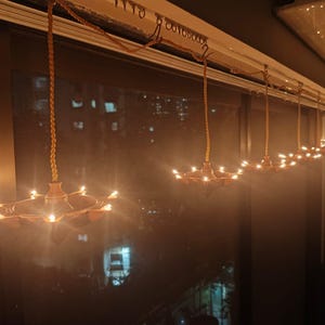 May include: A series of hanging oil lamps with lit wicks, suspended from a rope. The lamps are bronze-coloured and have multiple receptacles for the wicks. The background is dark, with a window showing city lights.