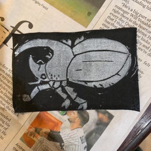 May include: A black fabric patch with a white outline of a beetle with large horns. The beetle is facing the right side of the image.
