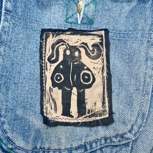 Op de afbeelding: Een handgeprint ontwerp in zwart en wit van een wezen met grote ogen en een rond lichaam, genaaid op een blauw denim jack.
