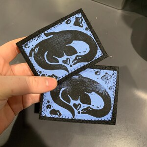 Könnte beinhalten: Zwei blaue und schwarze Filzpatches mit einer schwarzen Silhouette einer Meerestierfigur auf jedem. Die Patches sind mit schwarzem Garn um die Ränder genäht.