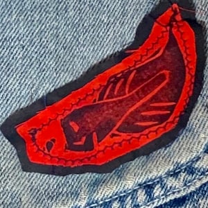 May include: Red and black fabric patch with a stylized design sewn onto a blue denim background.