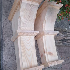 Puede incluir: Dos ménsulas de madera sin terminar, con una veta de madera clara y natural. Cada ménsula tiene una base rectangular, una columna central y una parte superior decorativa. Las ménsulas están diseñadas para soporte arquitectónico.