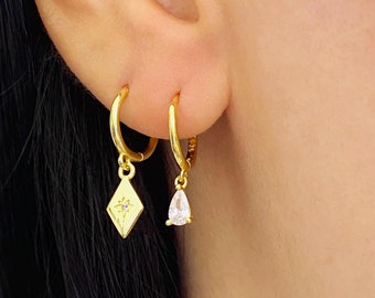 May include: A pair of gold hoop earrings with a small diamond-shaped charm and a teardrop-shaped charm with a clear stone.