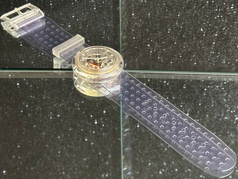 以下が含まれることがあります： 透明なプラスチック製の腕時計で、丸い文字盤と透明なプラスチック製のバンドが付いています。時計の文字盤は銀色の文字盤で、黒い目盛りと針が付いています。