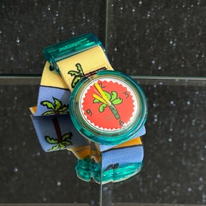 Puede incluir: Un reloj de pulsera azul turquesa con un diseño de palmera roja y verde en la esfera. El reloj tiene una banda de tela a rayas amarillas y azules.