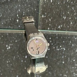 May include: A silver Swatch watch with a black strap and a gray face. The face has a unique design with a large number 5 and a smaller number 6. The watch has a silver second hand and a gold hour hand.