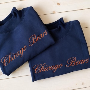 May include: Two navy blue sweatshirts featuring a mock neck collar and the text Chicago Bears embroidered in orange cursive script across the front. The garments are folded and layered, showcasing the dark blue fabric and the orange lettering.