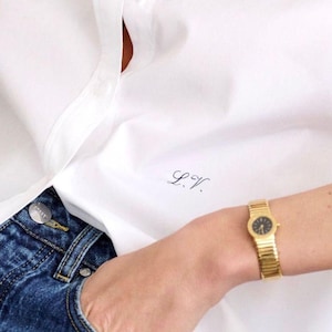 May include: A white button-down shirt with the initials "L.V." embroidered on the front. The shirt is paired with blue jeans and a gold watch with a black face. A gold chain necklace is also visible.