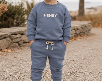 May include: A dusty blue long-sleeve sweatshirt and matching sweatpants set. The sweatshirt features the name HENRY printed in white capital letters on the chest. The sweatpants have a white drawstring at the waist and ribbed cuffs.