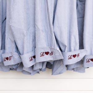 May include: Several blue and white striped dress shirts with embroidered initials and hearts on the cuffs. The shirts are hanging against a white wooden background.