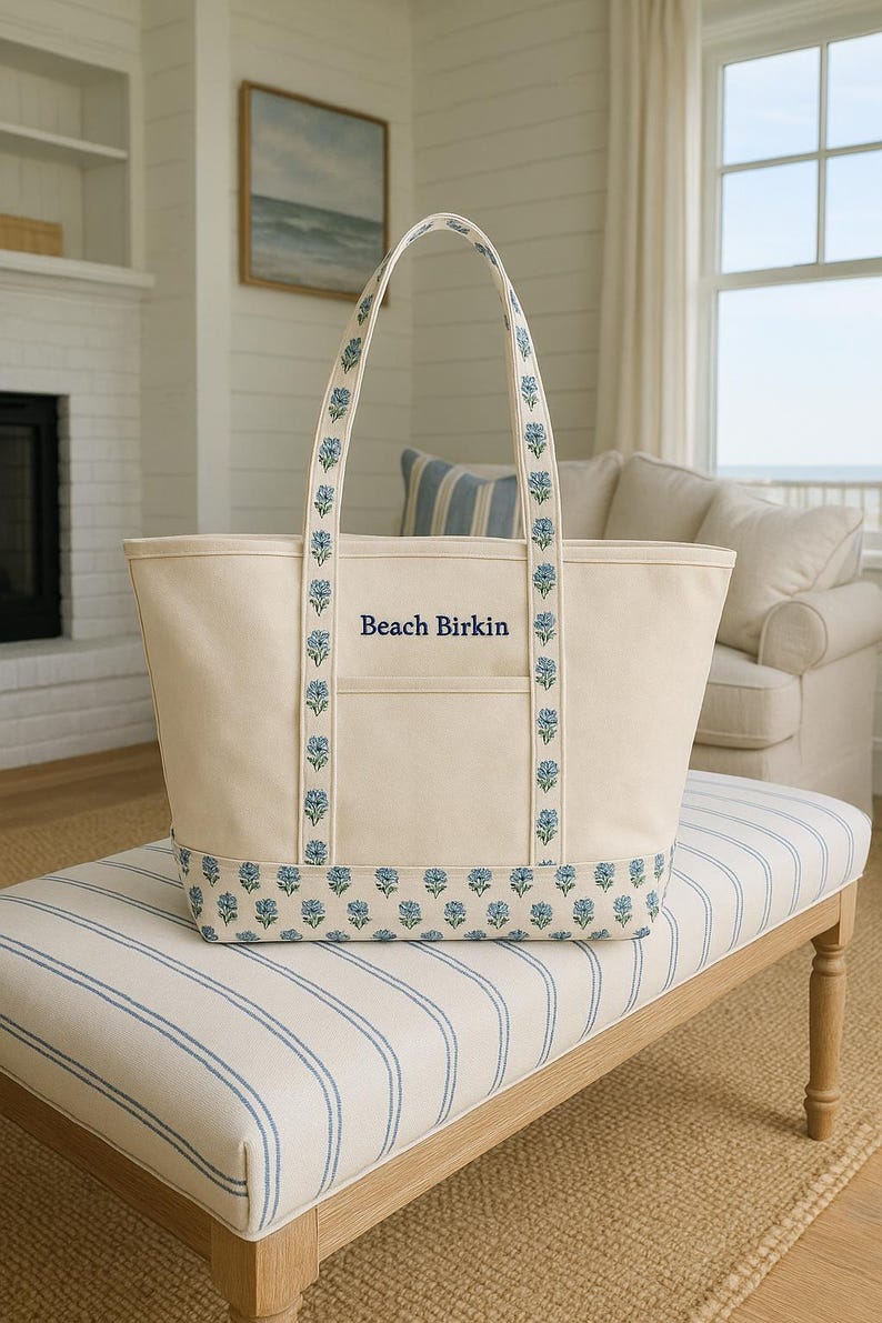 May include: Cream-colored tote bag with blue floral accents and the text "Beach Birkin." The bag features blue floral patterns on the handles and base. It is sitting on a bench with blue and white stripes.