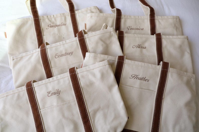 May include: Several off-white canvas tote bags with brown handles and trim are arranged on a white surface. Each bag has a name embroidered on the front: Jacqueline, Courtney, Emily, Veronica, Alissa, and Heather.
