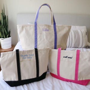 May include: Three canvas tote bags in varying sizes. The largest bag has purple handles and the name "Lila" embroidered on it. The other two bags have black and pink accents, with the phrases "Thank God for July" and "well read" embroidered on them.