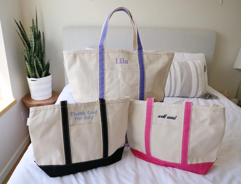 May include: Three canvas tote bags in varying sizes. The largest bag has purple handles and the name "Lila" embroidered on it. The other two bags have black and pink accents, with the phrases "Thank God for July" and "well read" embroidered on them.